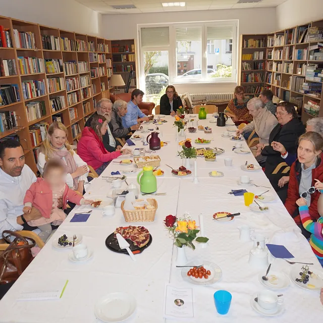 Erwachsene und Kinder sitzen um einen Tisch mit Brot, Butter, Käse, Weintrauben und Kuchen.